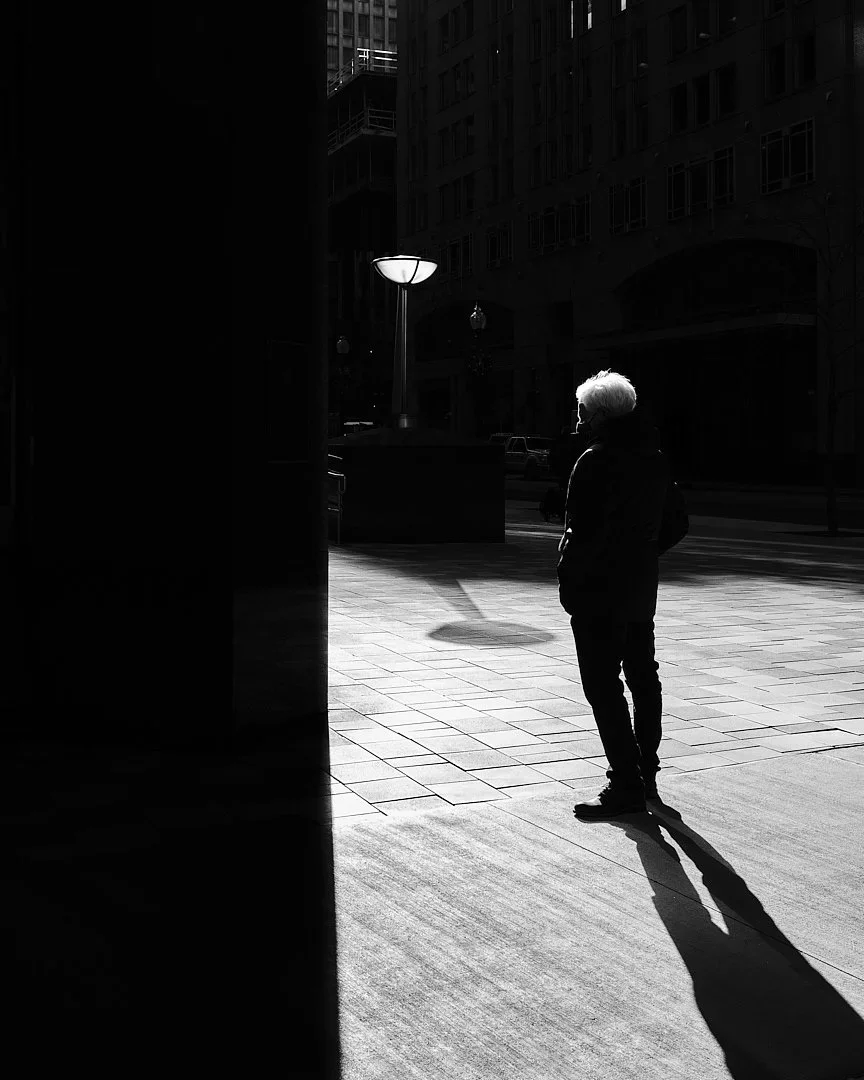 A man standing in a slice of sunlight on a dark street, casting a long shadow