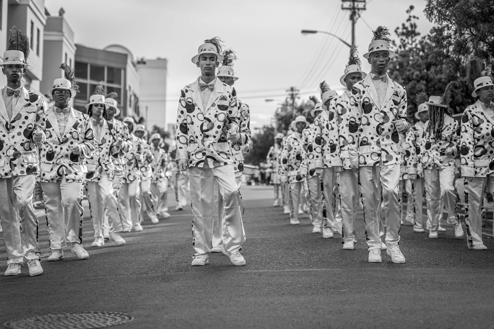 A Klopse troupe in matching patterned suits marching down the middle of a street toward the camera
