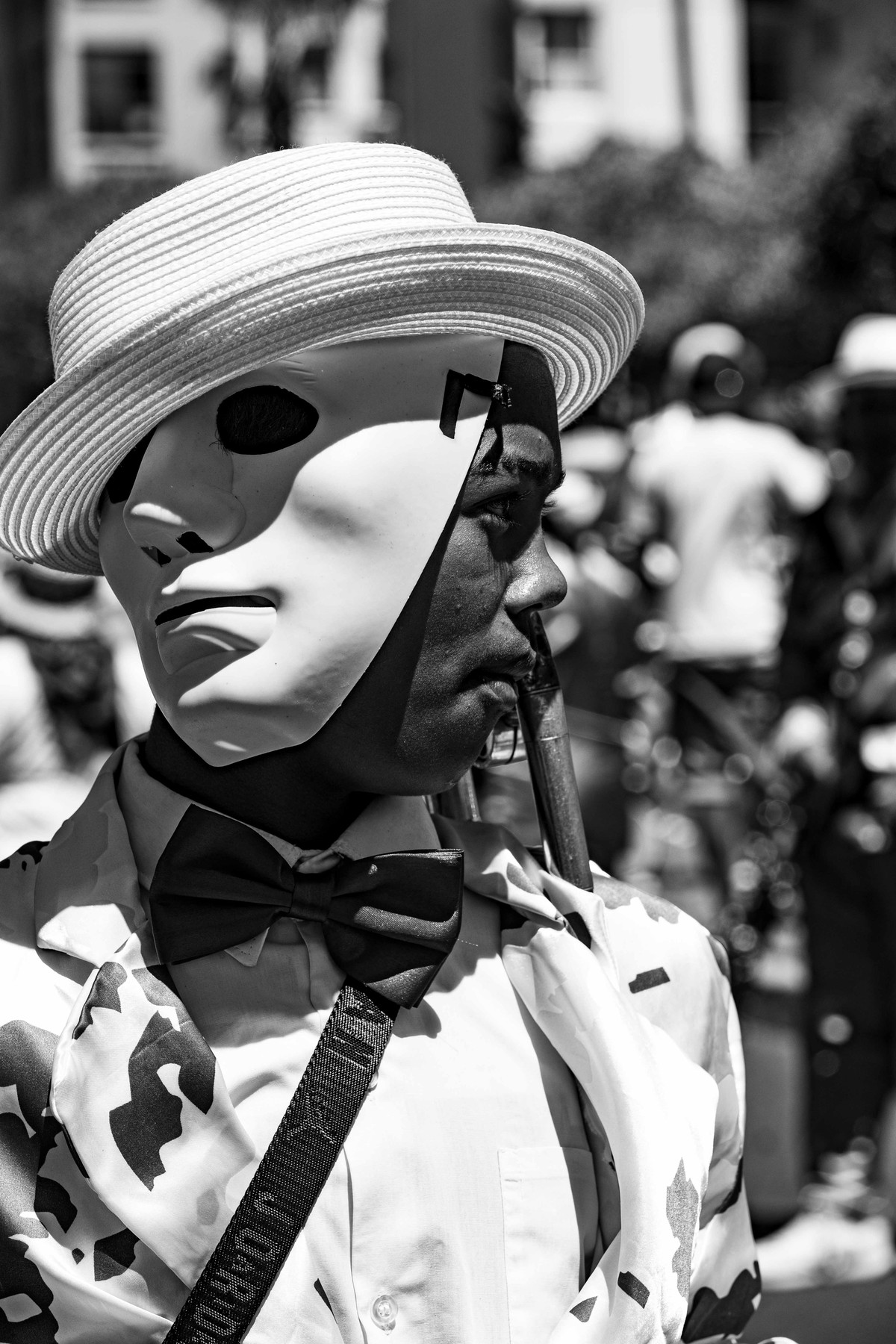 A performer in a white face mask and straw boater standing among the crowd