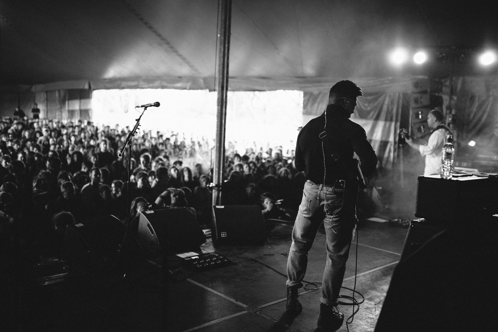 A guitarist seen from behind on a stage, dark against a bright tent opening and a large crowd