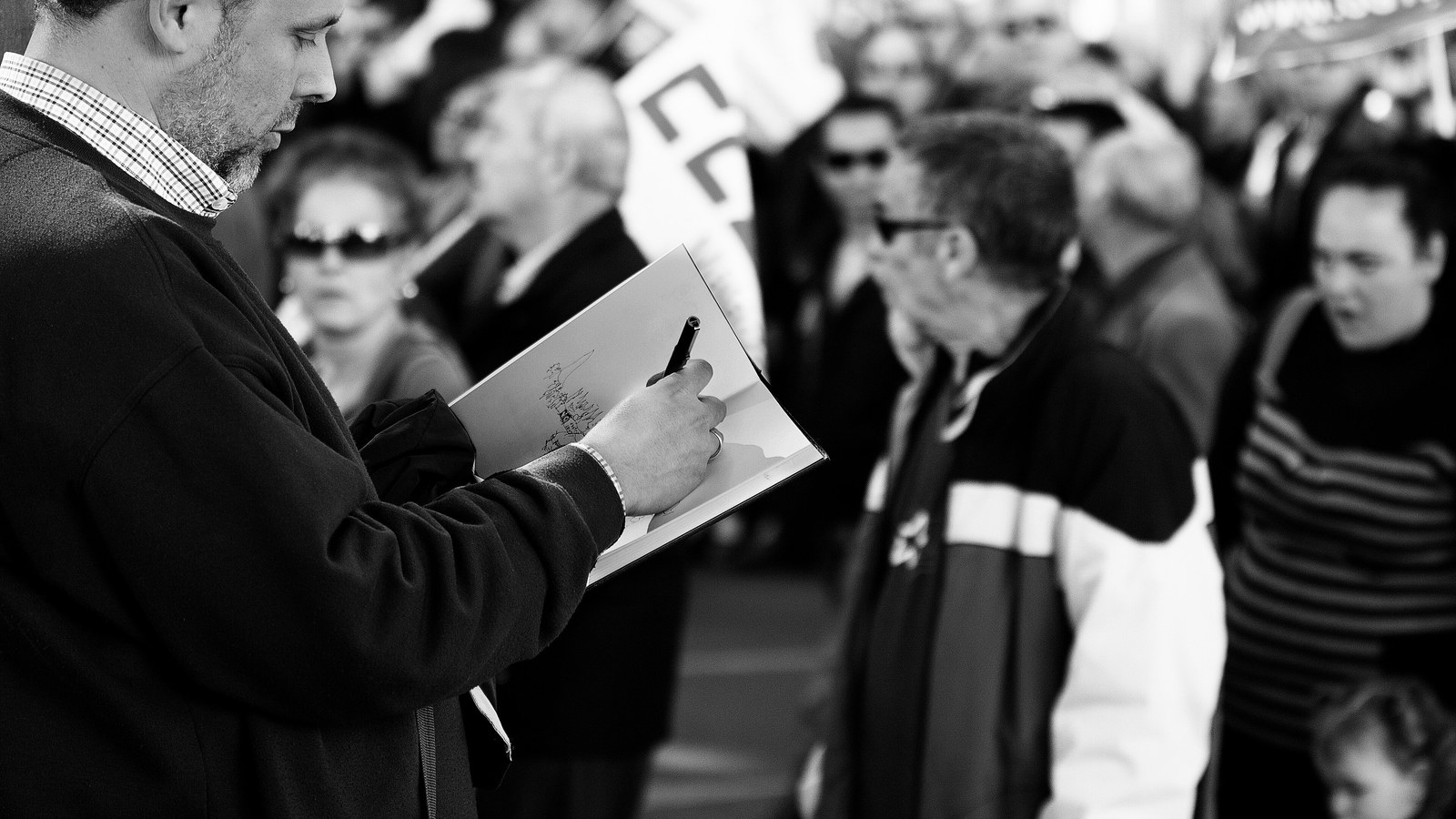 A man in the foreground drawing in a sketchbook while a dense, softly blurred crowd presses around him