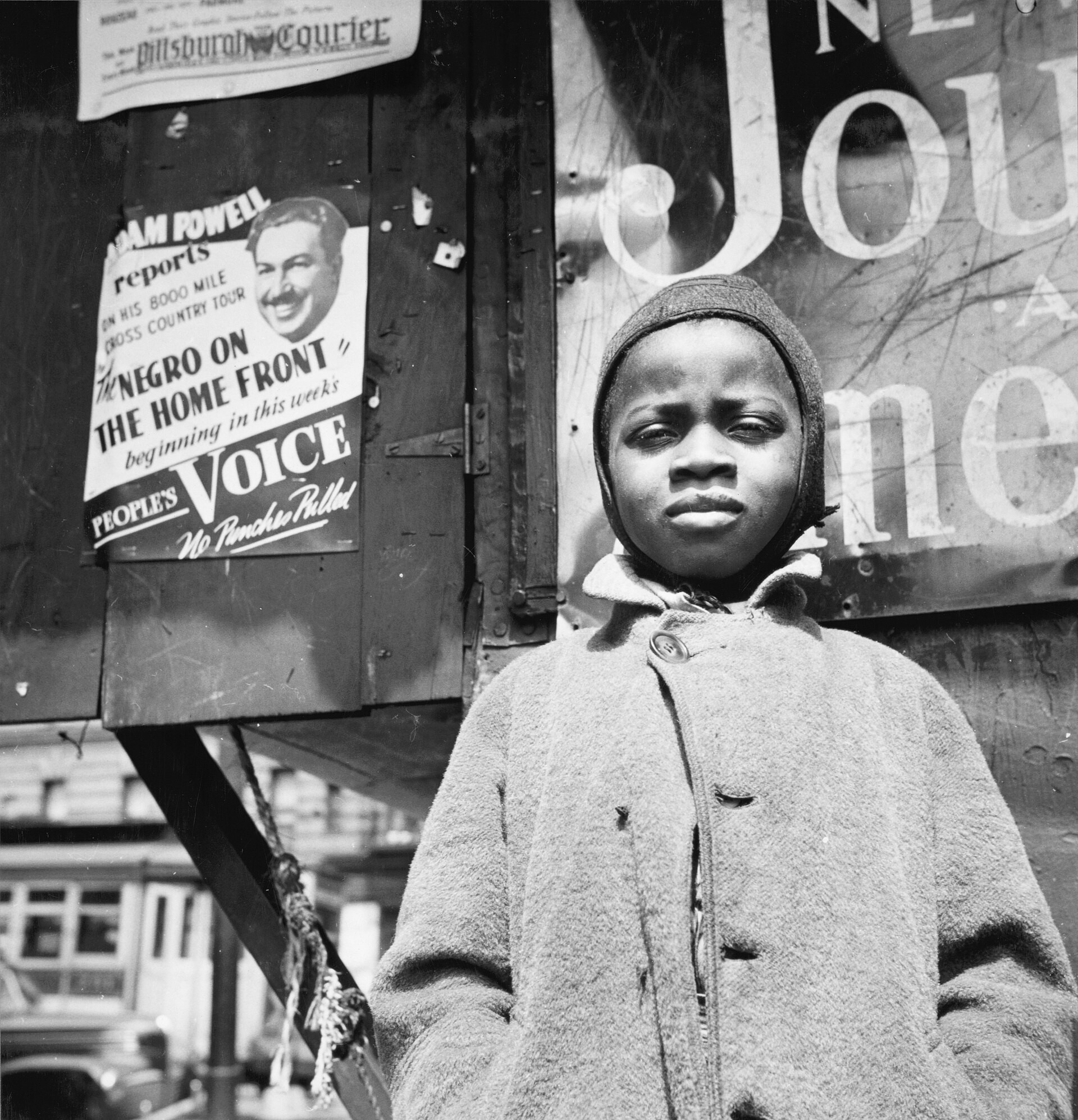A boy in a knitted hat and heavy coat standing in front of a Harlem newsstand covered in posters
