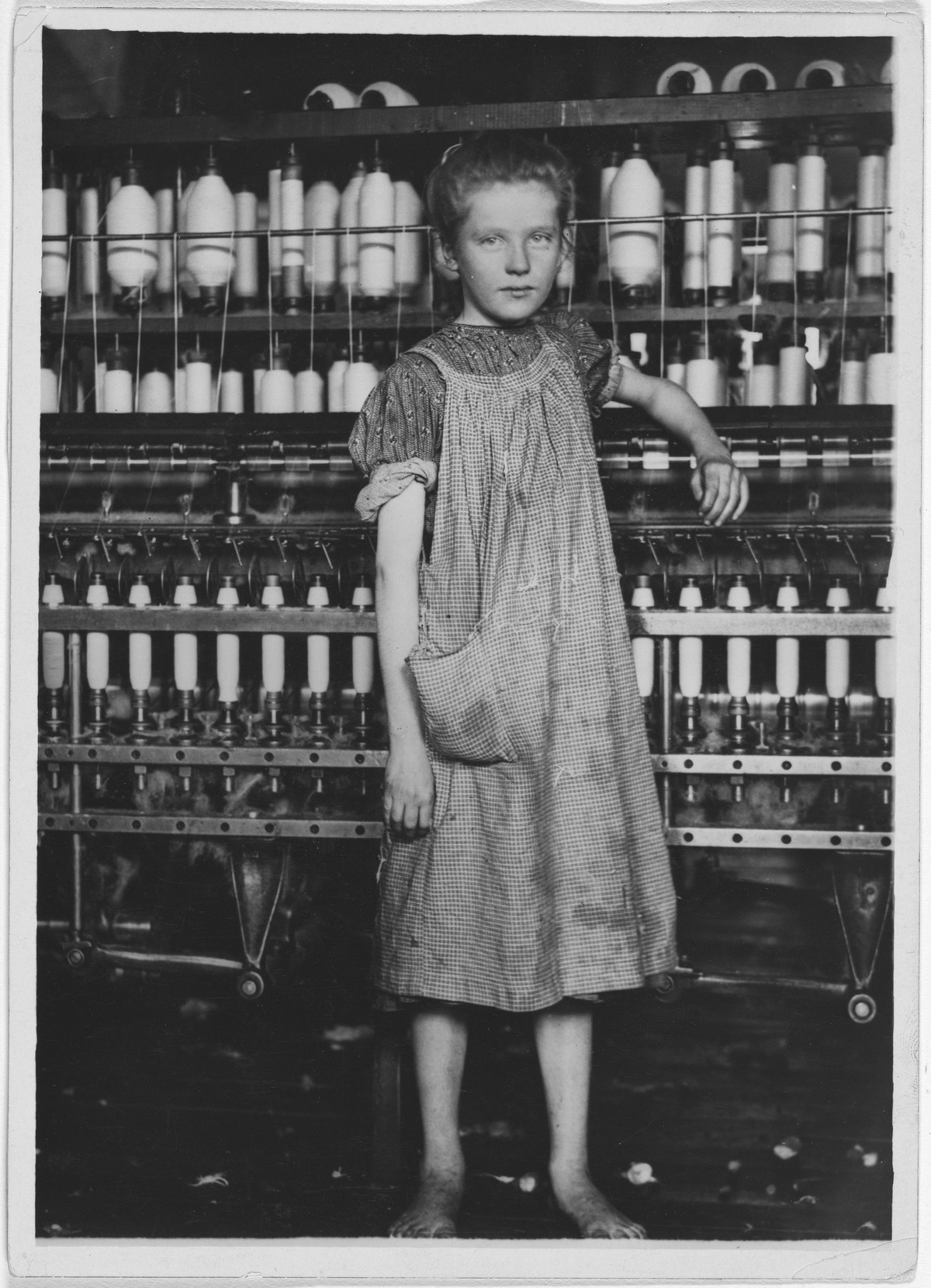 A barefoot girl in a checked dress leaning on a spinning frame in a cotton mill, looking at the camera