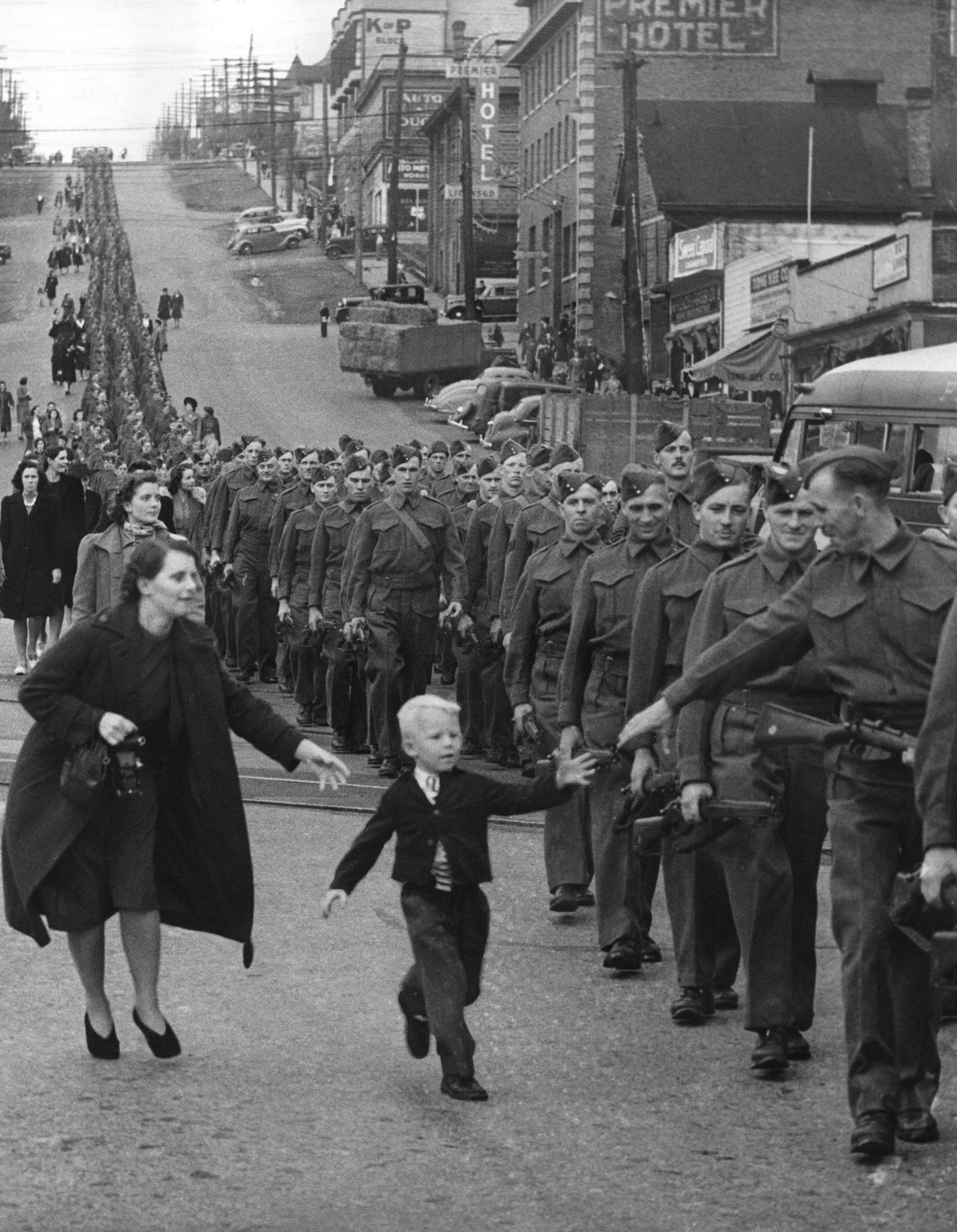 A small boy breaks from his mother and runs toward his father in a column of marching soldiers, his hand outstretched