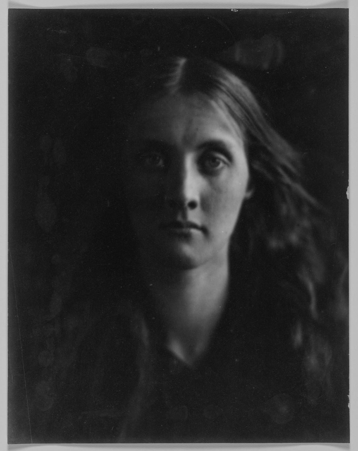 A close portrait of a young woman with long loose hair looking straight at the camera out of deep shadow