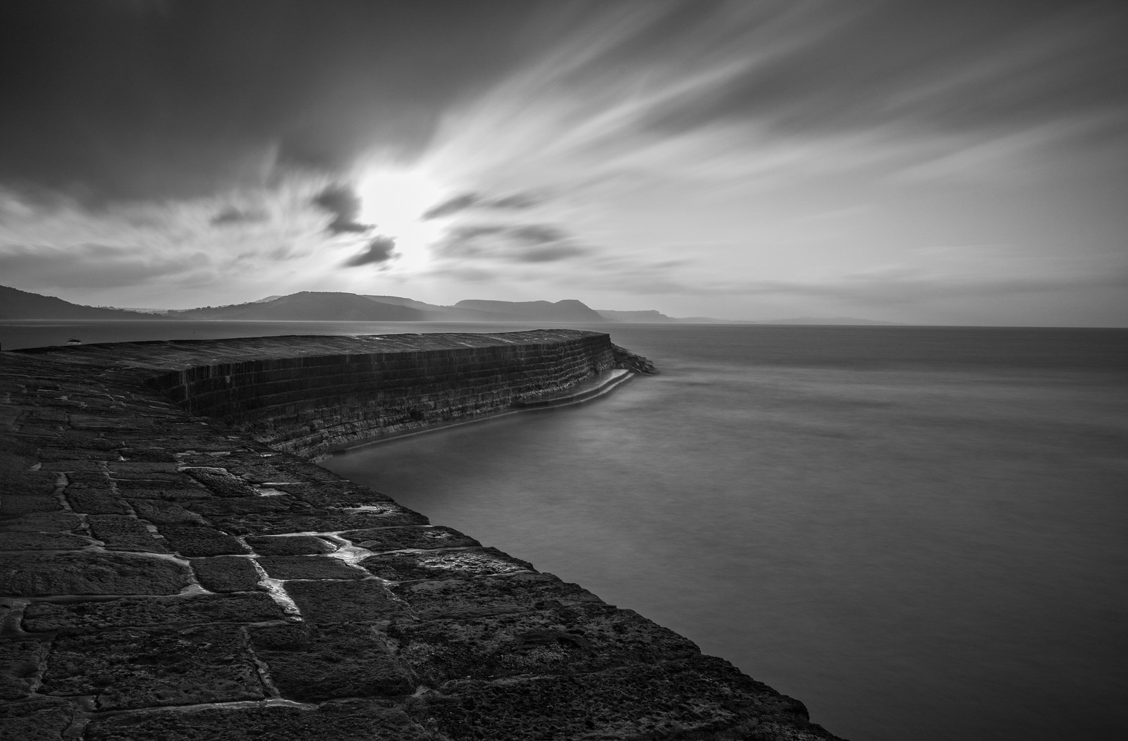A long exposure of a curving stone harbor wall at Lyme Regis, with smooth water and clouds drawn into streaks