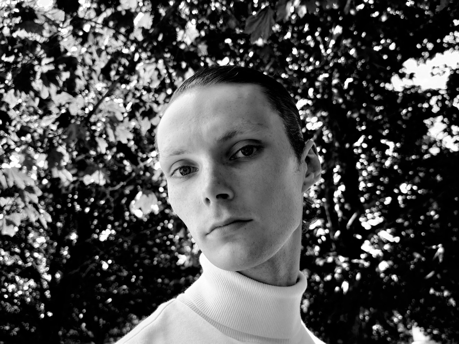 Black and white portrait of a person in a roll neck against a bright tree canopy thrown out of focus behind them