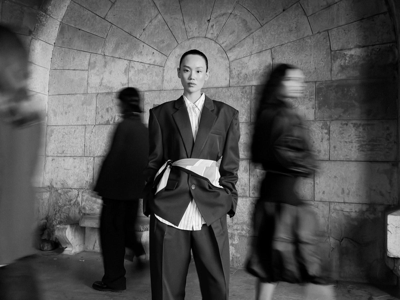 Black and white photograph of a person standing still under a stone arch while two people blur past on either side
