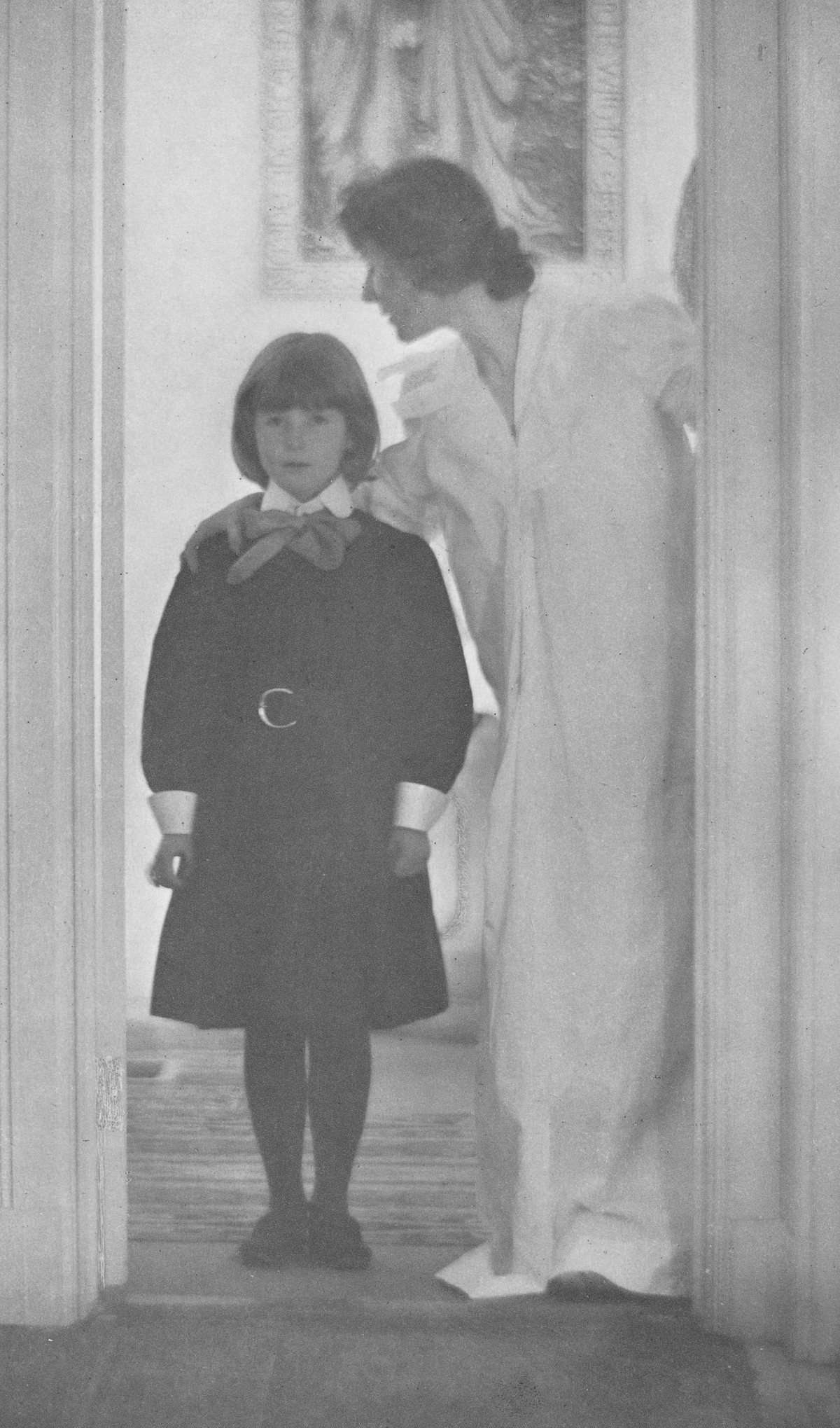 A woman in a long white gown leans toward a young girl in a dark dress standing in a doorway