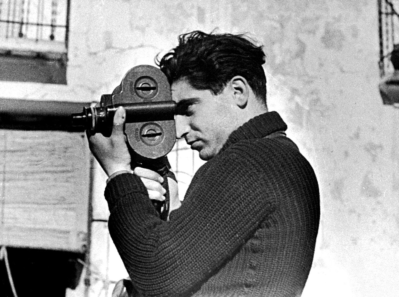 Robert Capa in a heavy knit sweater holding a film camera to his eye