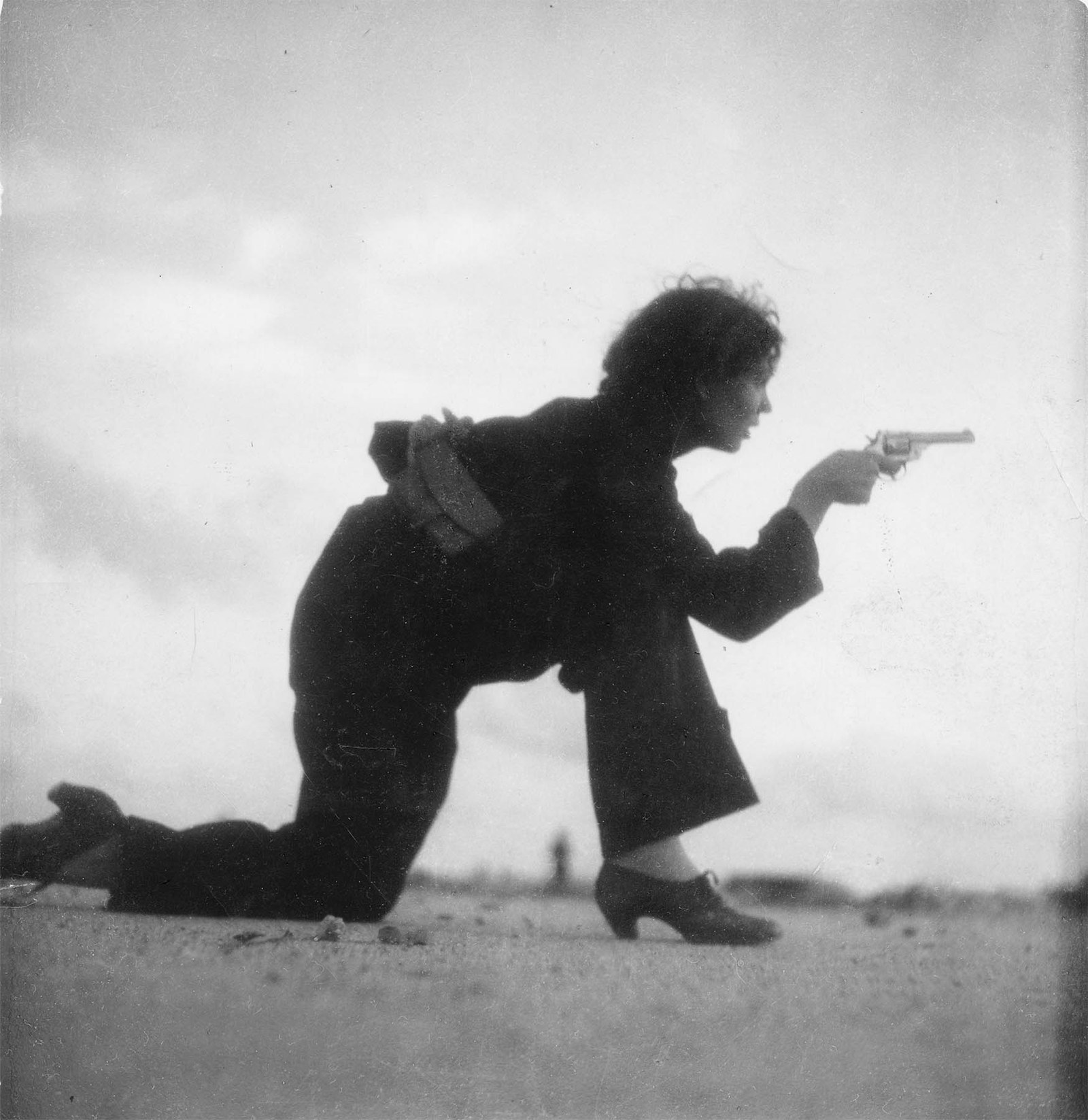 A woman in dark clothes kneeling on a beach and aiming a pistol, training for a Republican militia