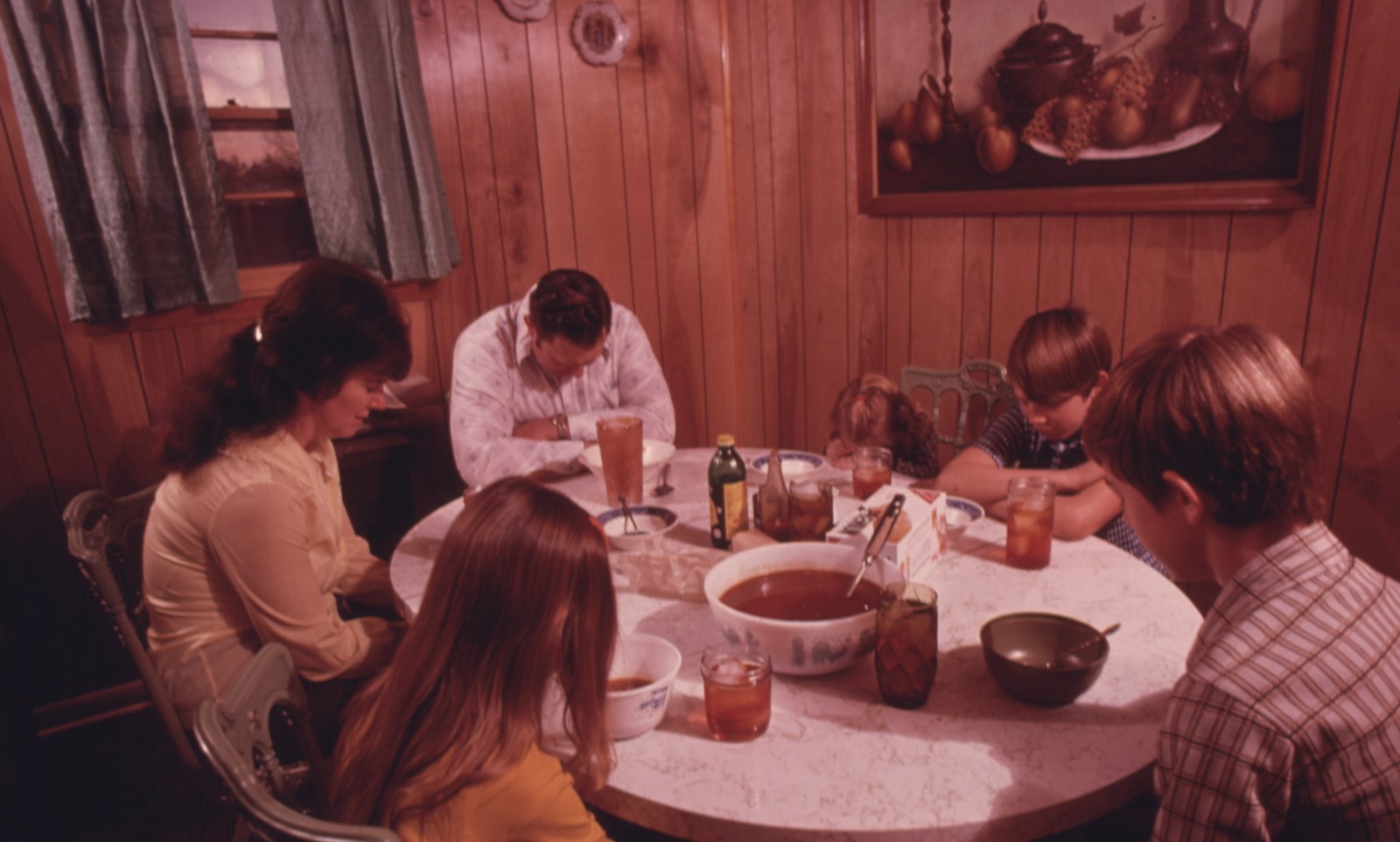 A family with four children bowing their heads in prayer around a kitchen table set with bowls and glasses of iced tea, in a room with wood paneled walls