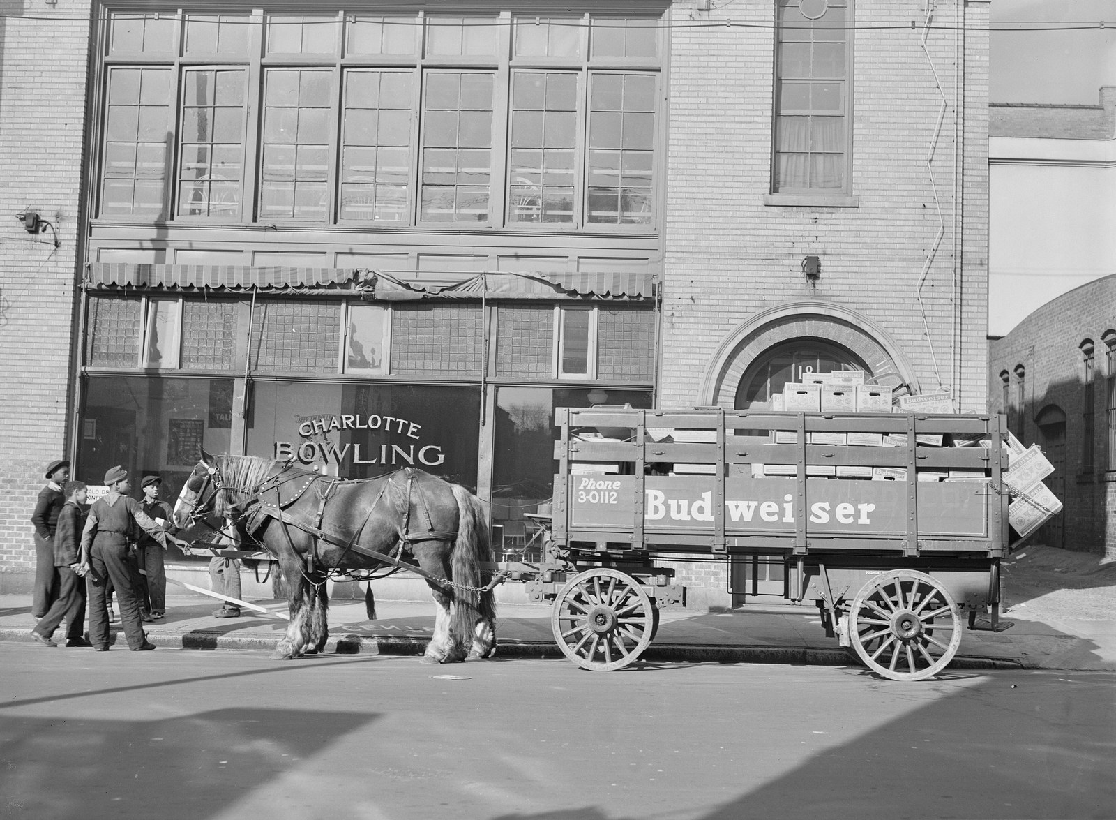 A horse drawn Budweiser delivery wagon parked outside a bowling alley, with young men standing by the horse