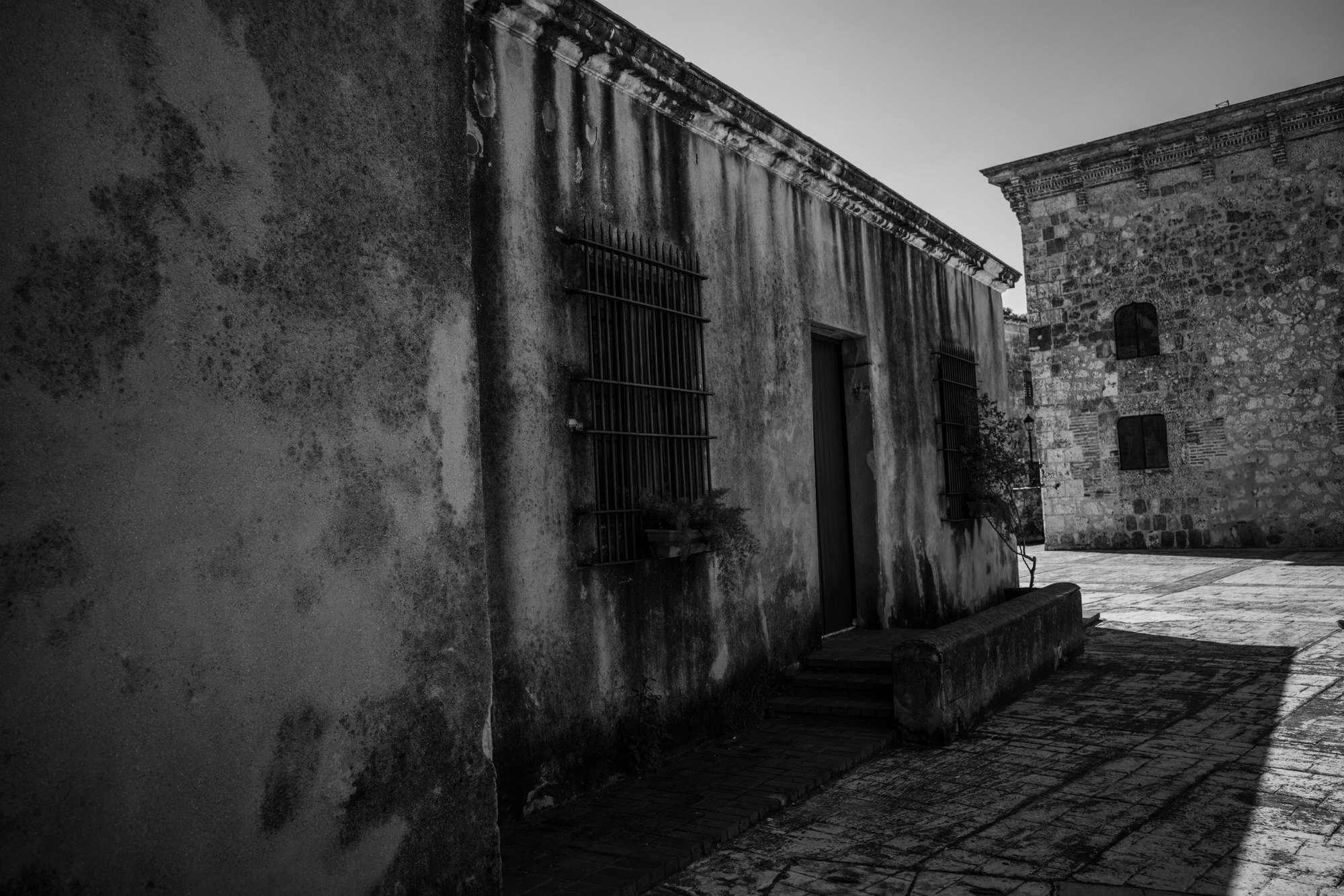 A weathered colonial wall with a heavy barred window, a narrow empty street running alongside it in hard sunlight