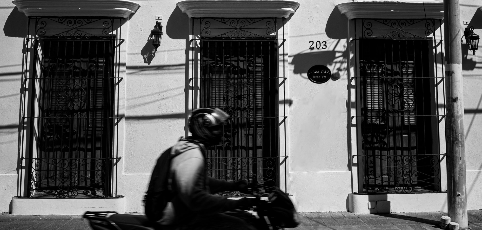 A rider on a motorcycle passing three barred windows set into a pale colonial wall in the Zona Colonial