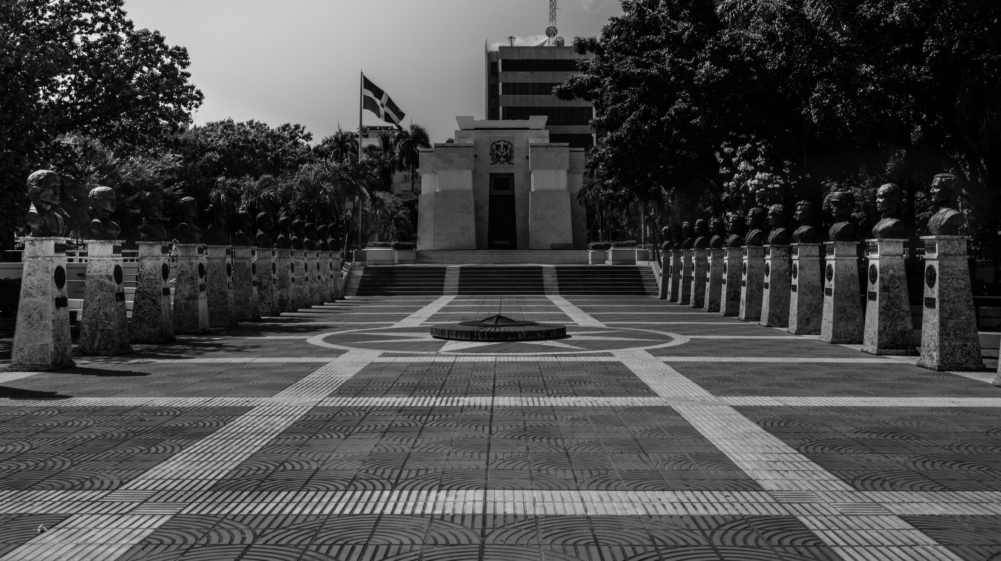 A long symmetrical walkway of patterned paving leading to a monument in Santo Domingo, with trees along both sides and a flag flying above
