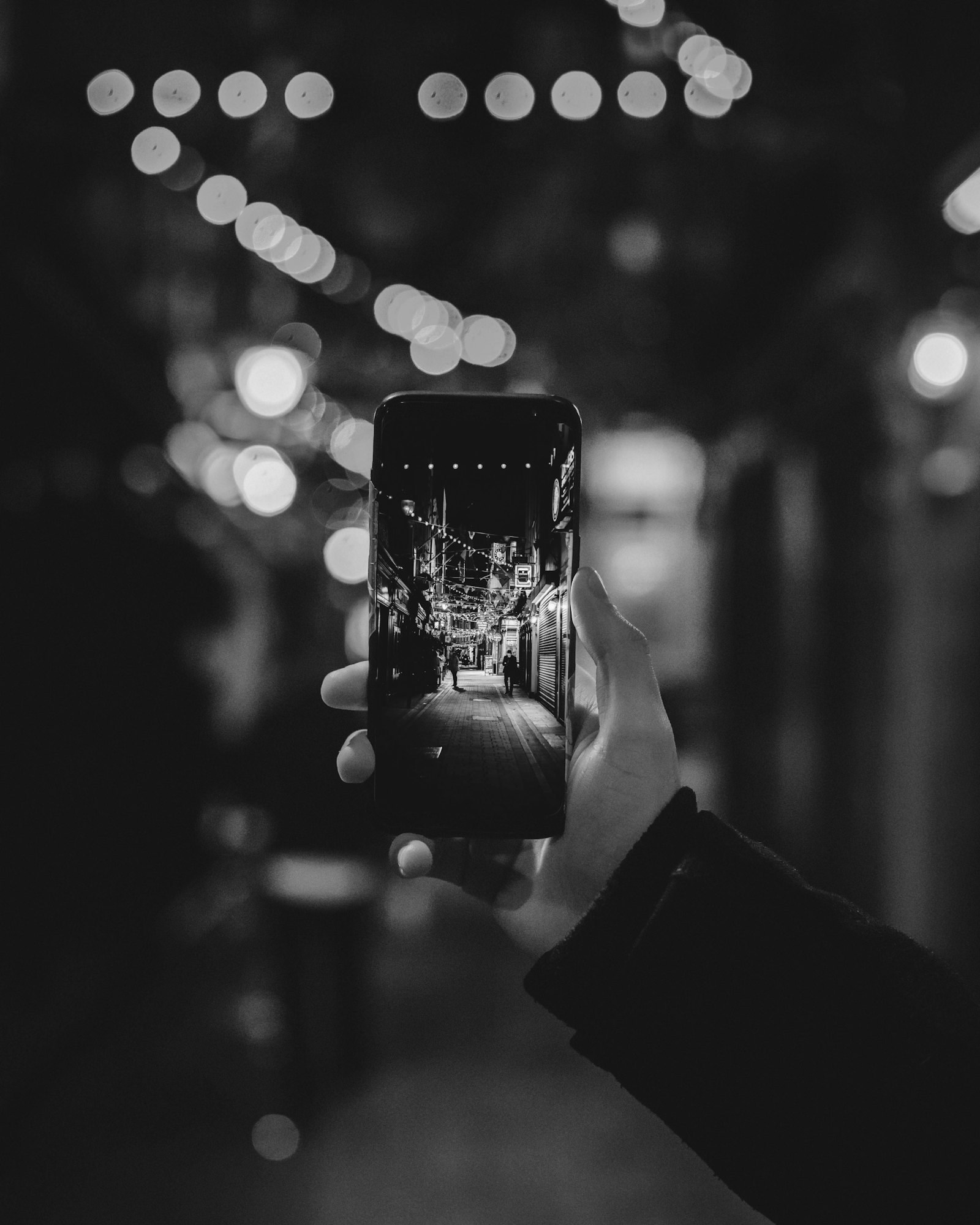 A hand holding a phone up to photograph a street at night, strings of lights going out of focus behind it