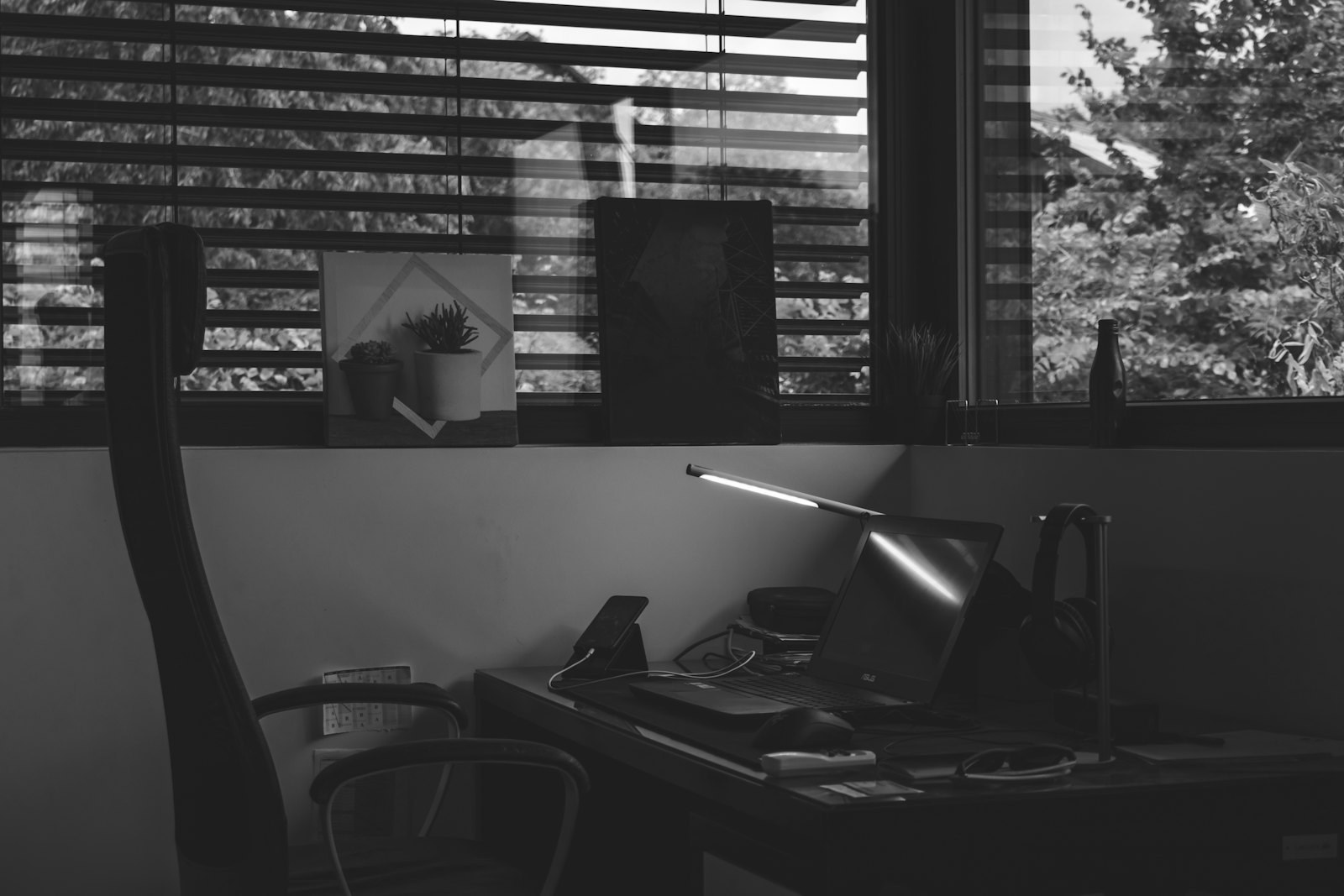 A cluttered desk lit by a single angled lamp, with window blinds and a chair pushed back
