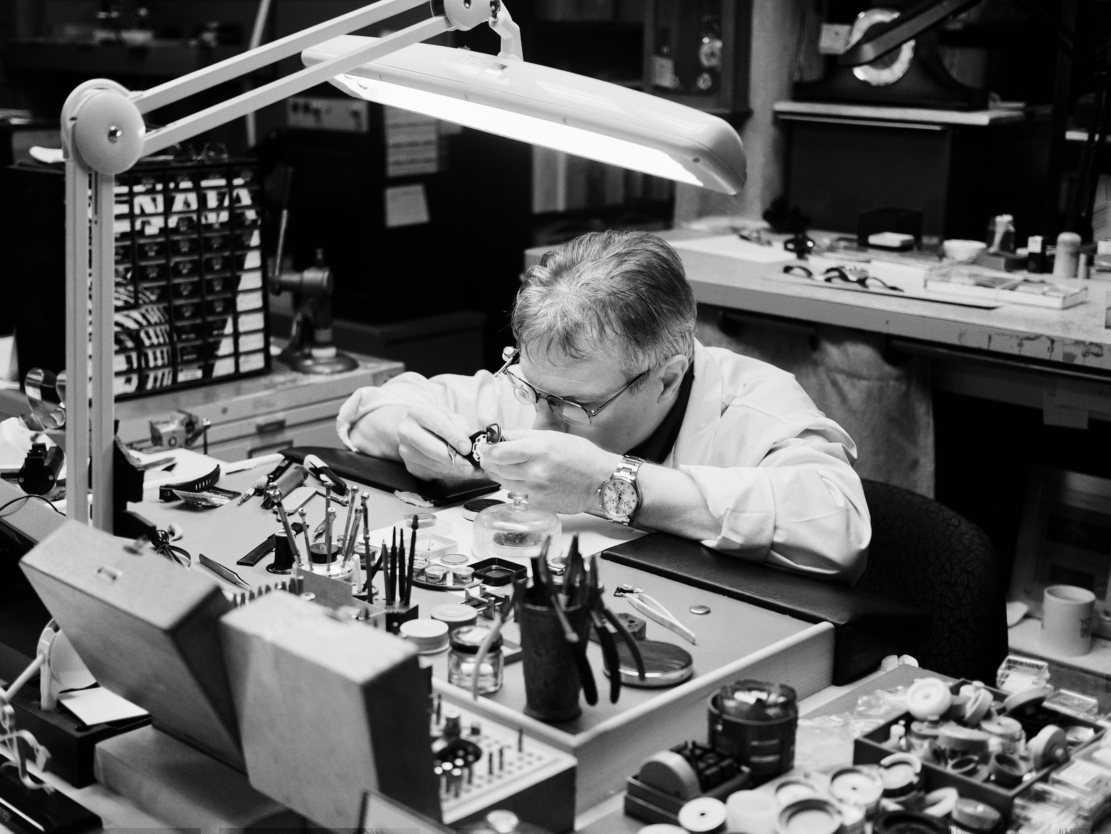 A watchmaker working under a bright lamp at a bench covered with small tools