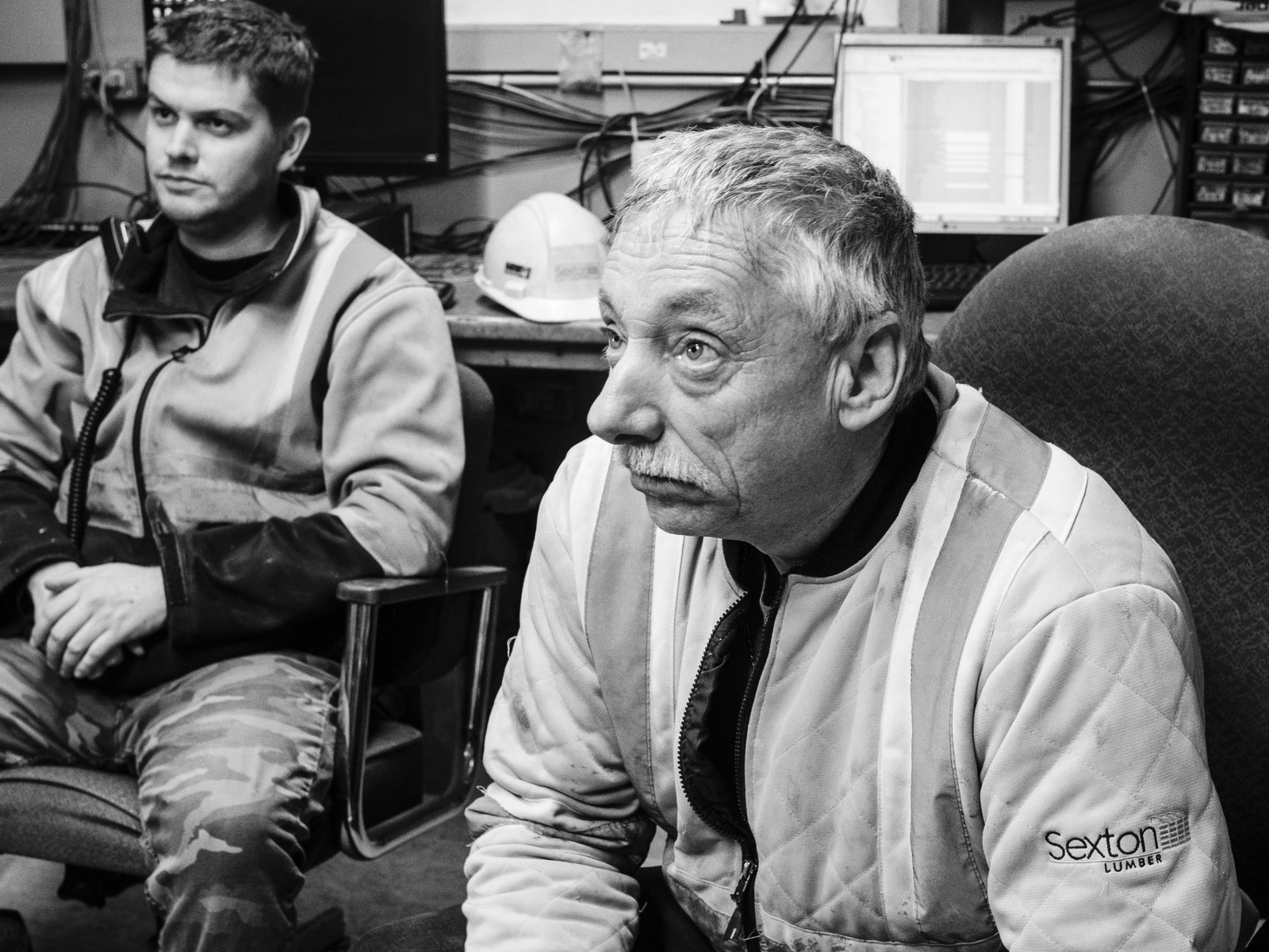 Two men in work jackets in a sawmill control room, screens behind them