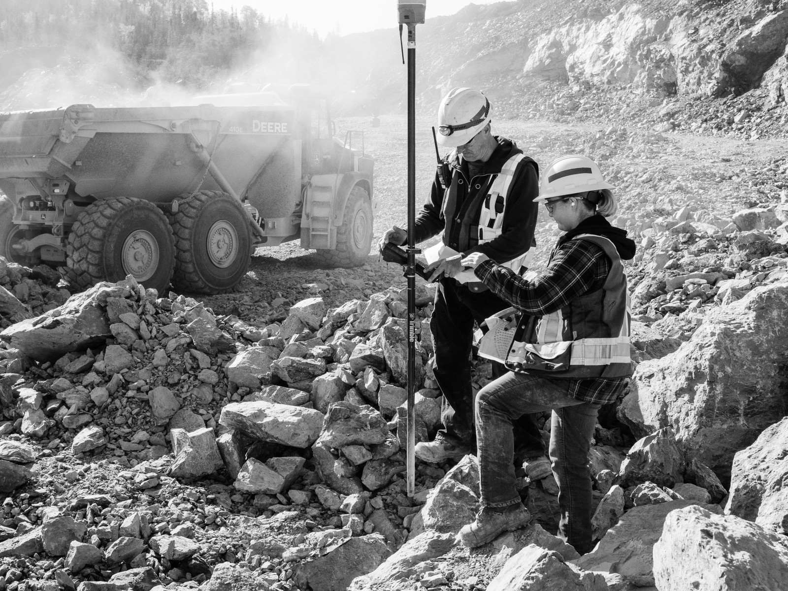Two surveyors with equipment standing among broken rock, a haul truck behind them
