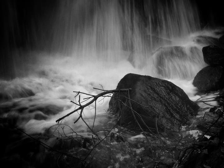 A long exposure of water falling past a dark boulder, with bare branches crossing the foreground