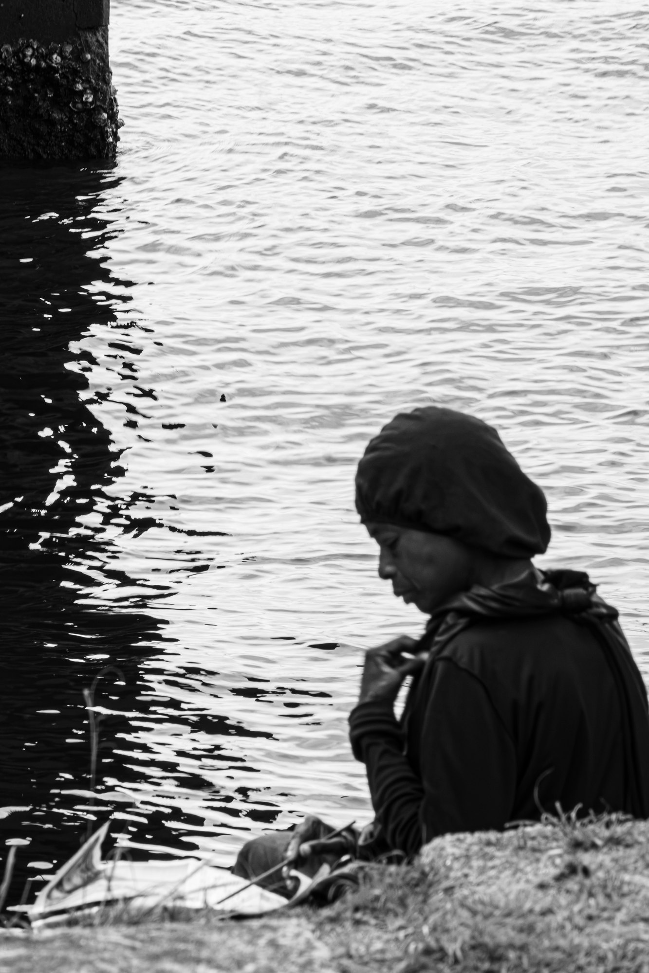 A woman in a headwrap sits on the grass at the waters edge working on a drawing