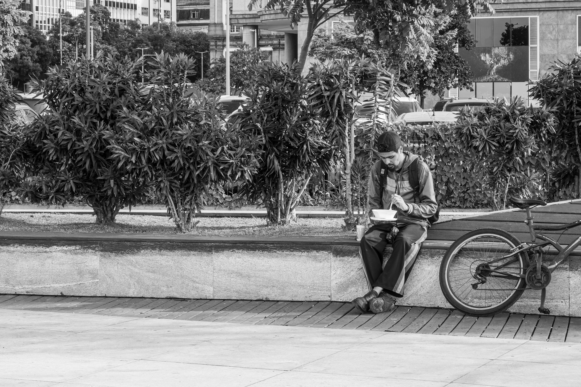 A delivery rider sits on a low wall eating from a lunch box with his bicycle leaning beside him
