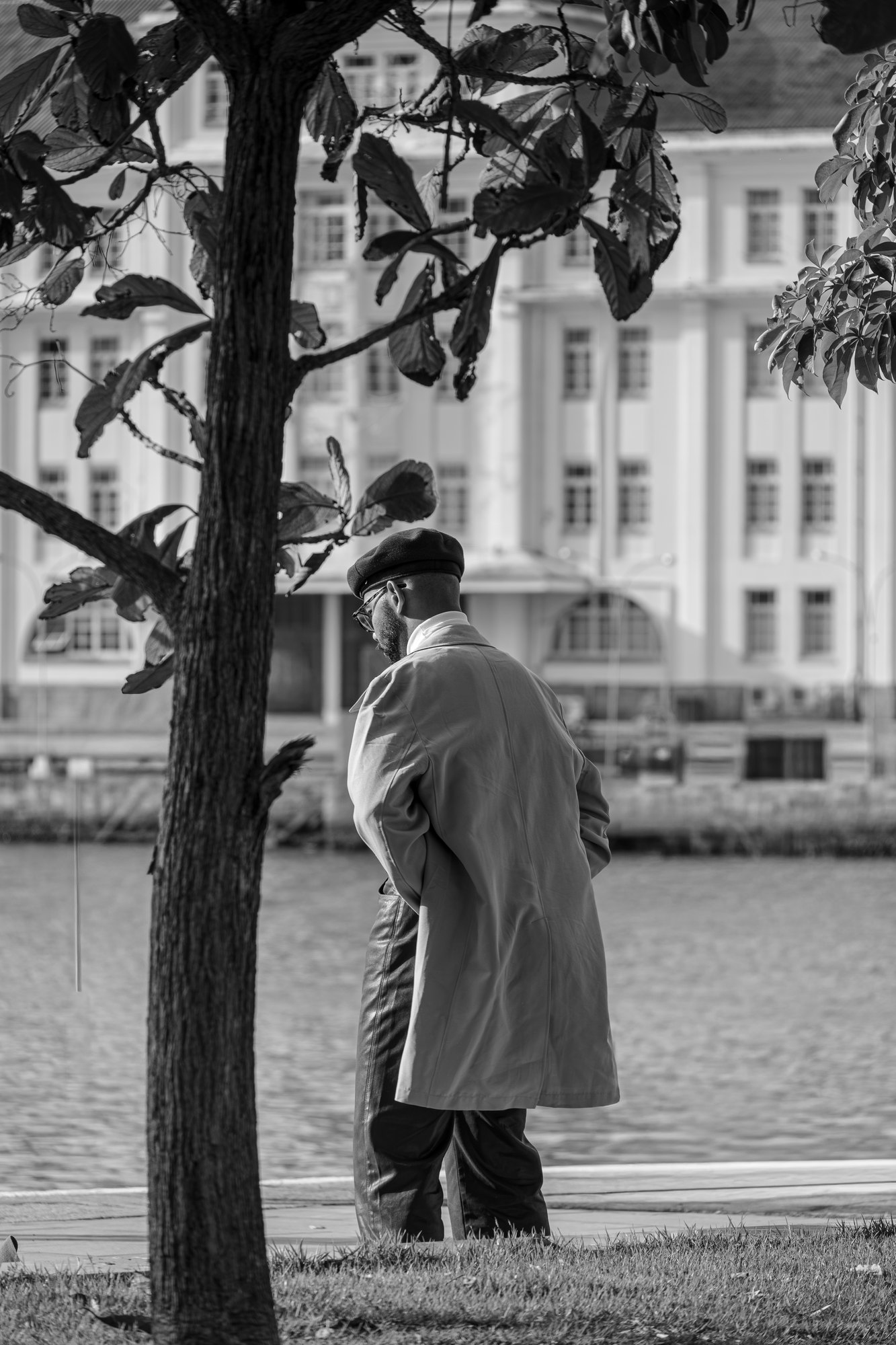 An older man in a long coat and flat cap walks beside the water in front of a pale colonial building