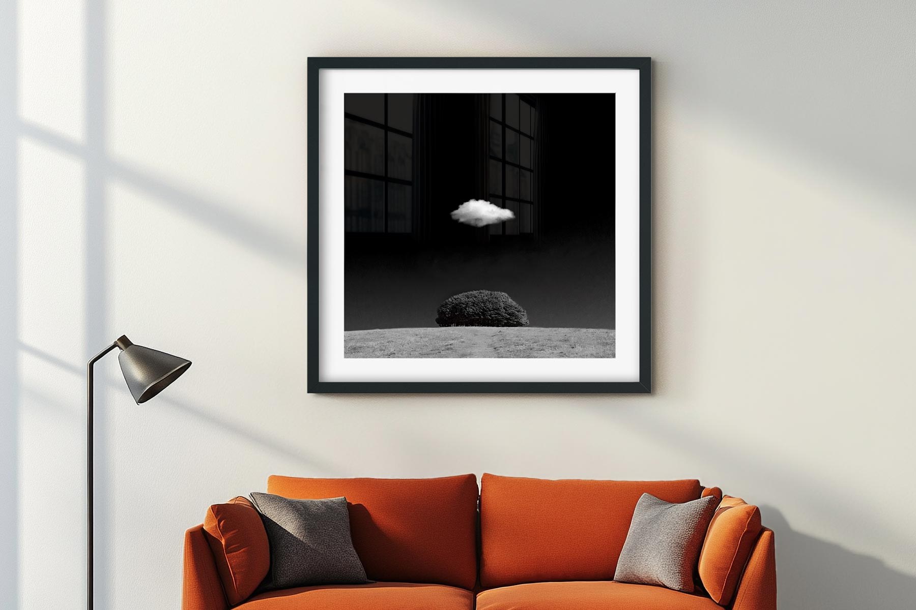 A framed print of a small dark tree on a hill beneath a single white cloud, hanging on a pale wall above an orange sofa
