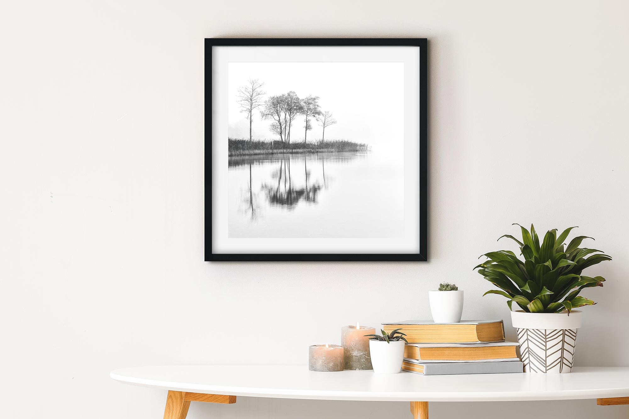 A framed print of trees reflected in still water, hanging above a white table with books and plants