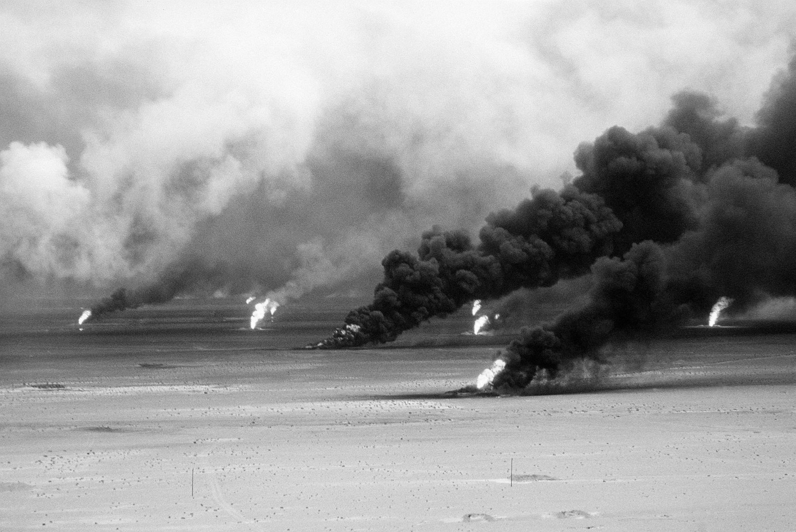 Oil well fires burning across the Kuwaiti desert in 1991, columns of black smoke rising from several wellheads across a flat pale plain
