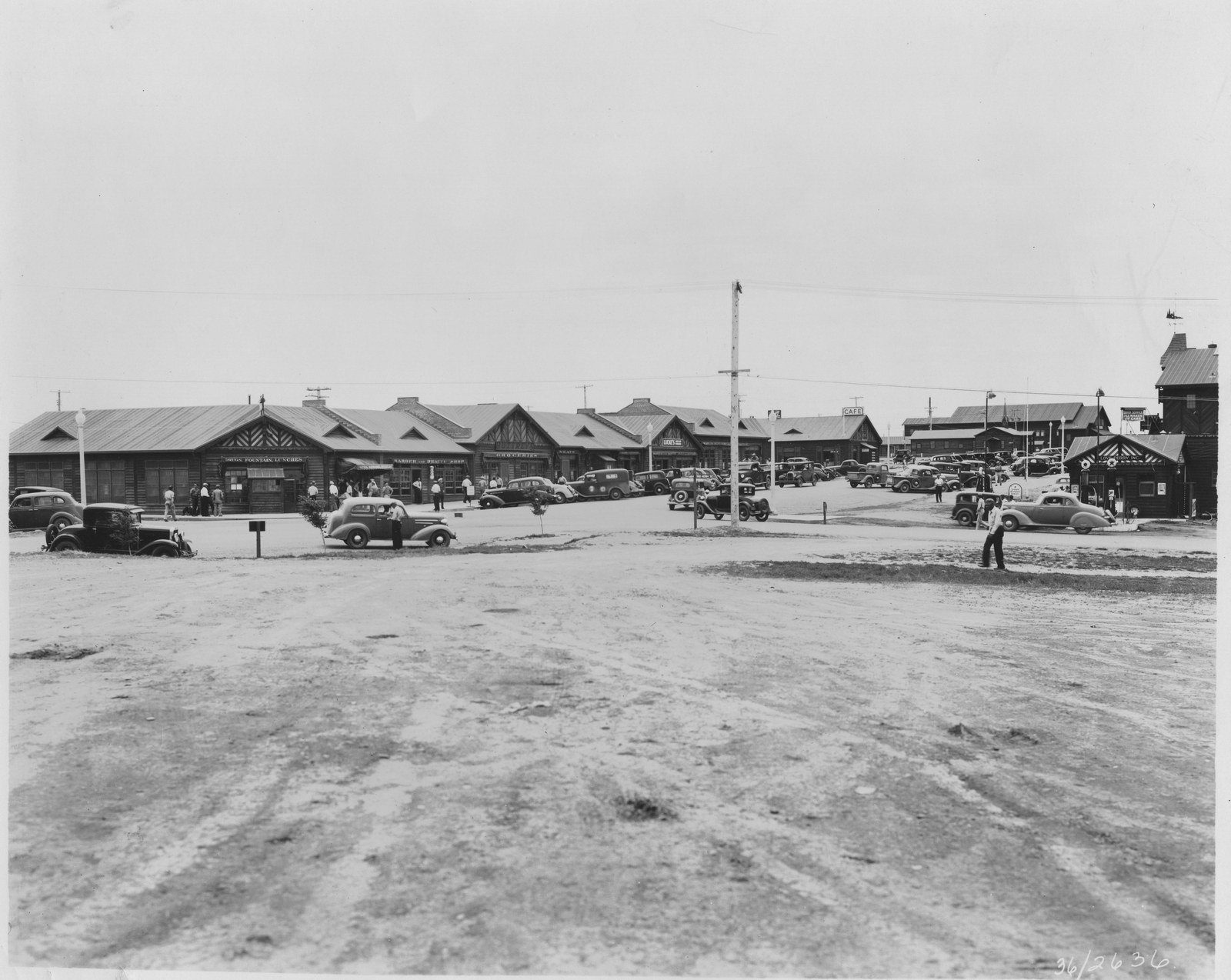 The commercial strip of the Fort Peck settlement in Montana in 1936, a row of wooden storefronts with a cafe, a barber shop and a soda fountain, with cars parked along a dirt road