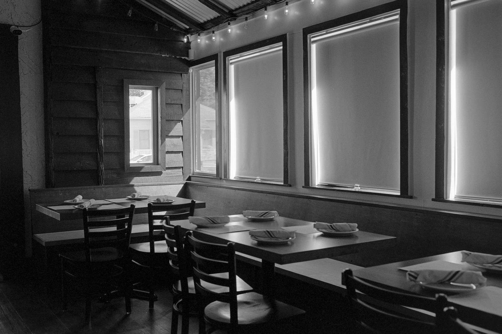 Empty tables and chairs in a restaurant dining room with sunlight burning around the edges of four drawn window shades