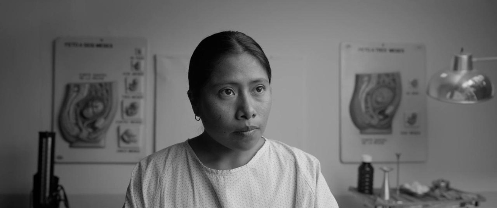 Cleo centered in a clinic, facing the camera, with matching anatomical charts hanging symmetrically on the wall behind her