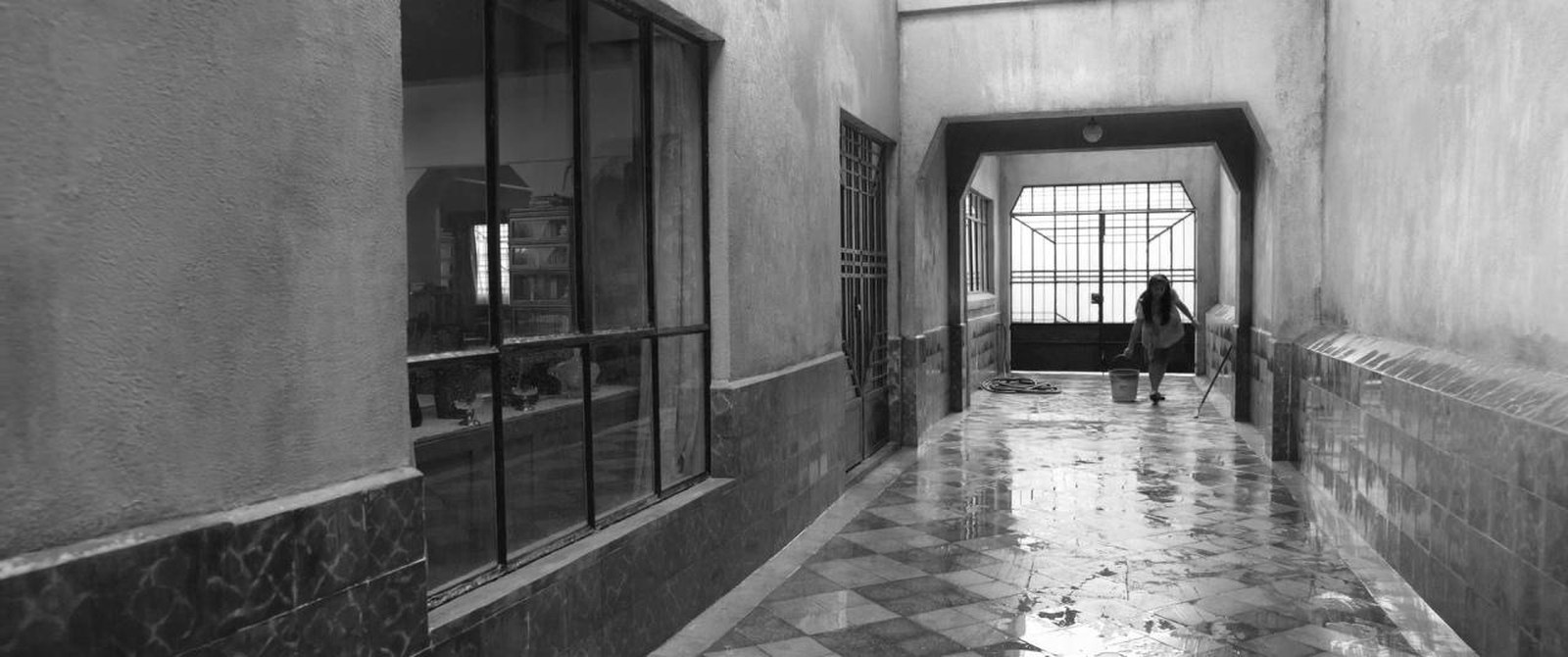 A long covered passage with a wet tiled floor and windows along one side, leading to a bright doorway with a small figure at the far end