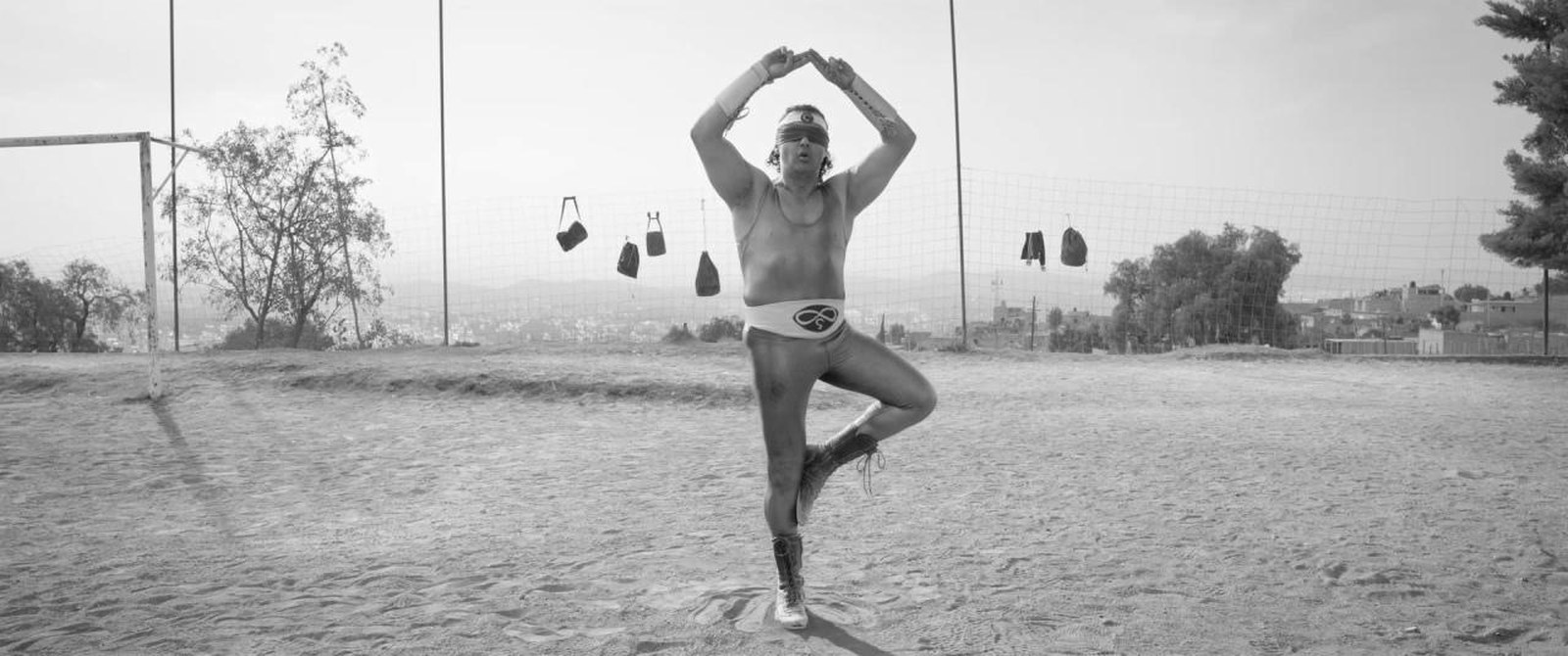 A lone man standing on one leg with his arms raised above his head in an open dirt field under a pale empty sky