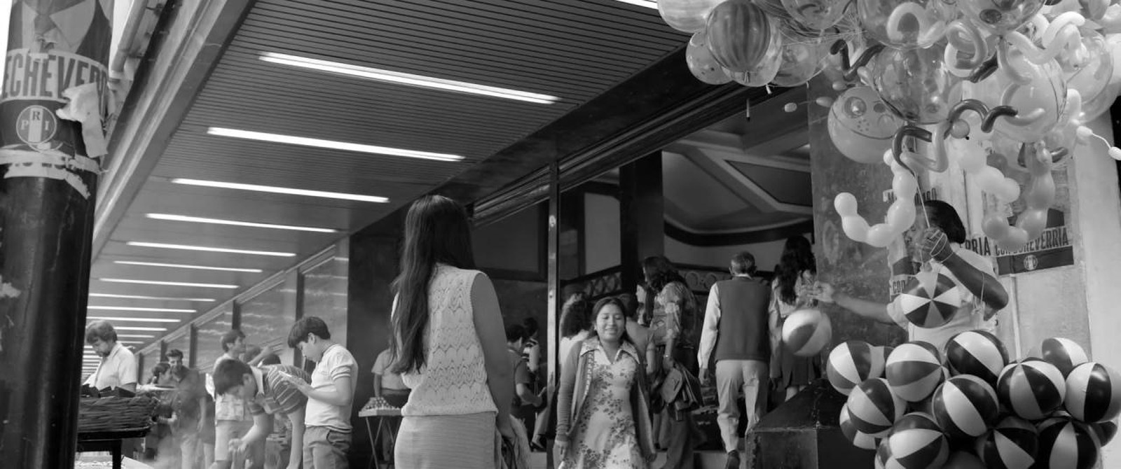 A crowded department store interior with a pillar in the foreground, a woman standing in the middle distance and shoppers and hanging beach balls behind her
