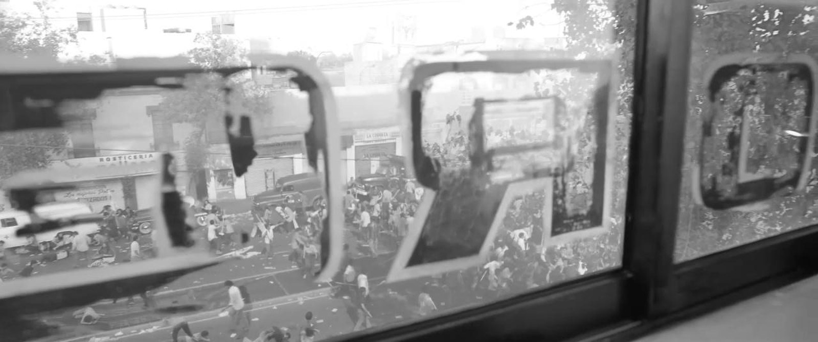 A view through a shop window with reversed painted lettering on the glass, looking out at a crowd in the street beyond