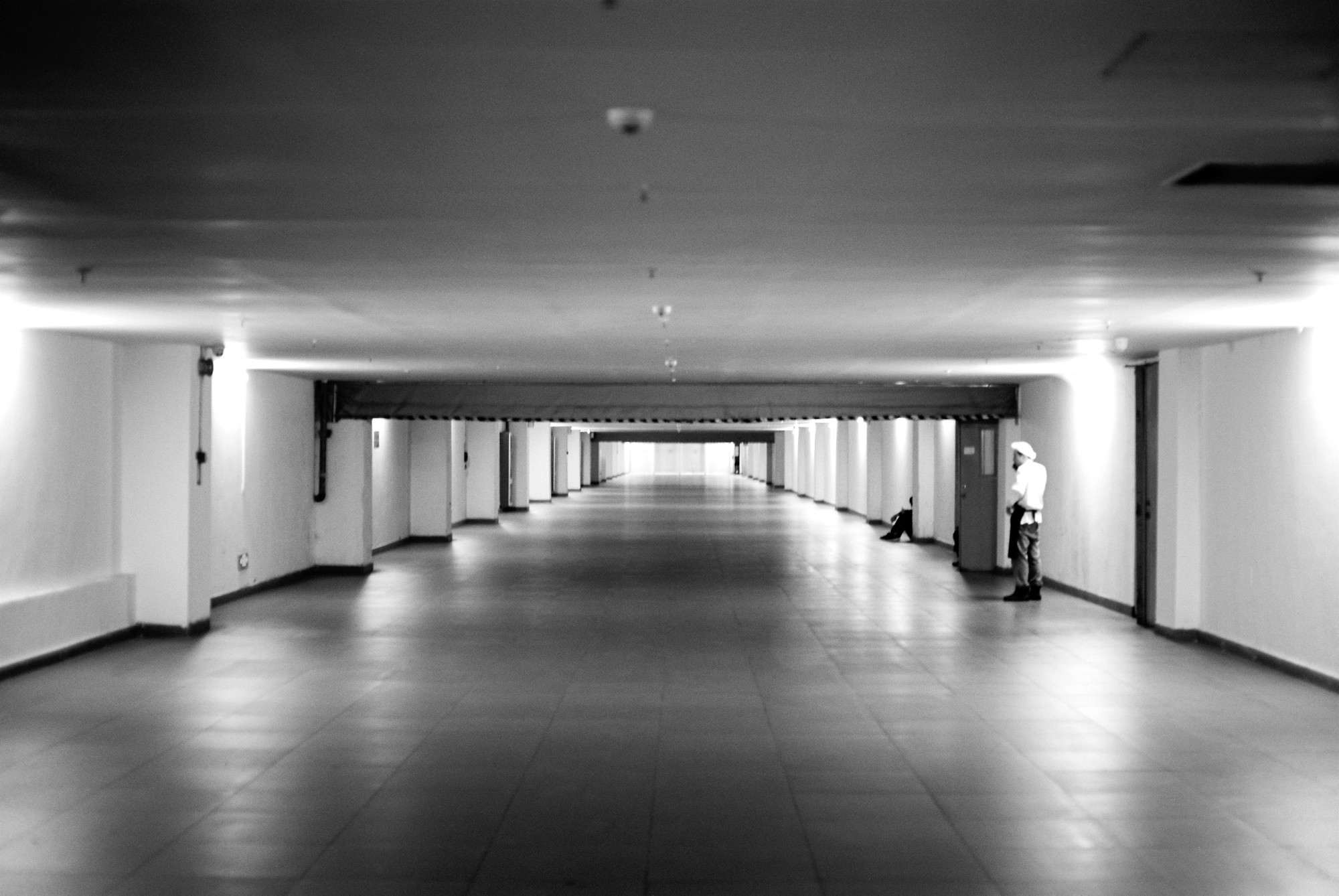 A long underground corridor photographed straight down the middle, repeating lights and columns receding into the distance, with one small figure standing at the right hand wall