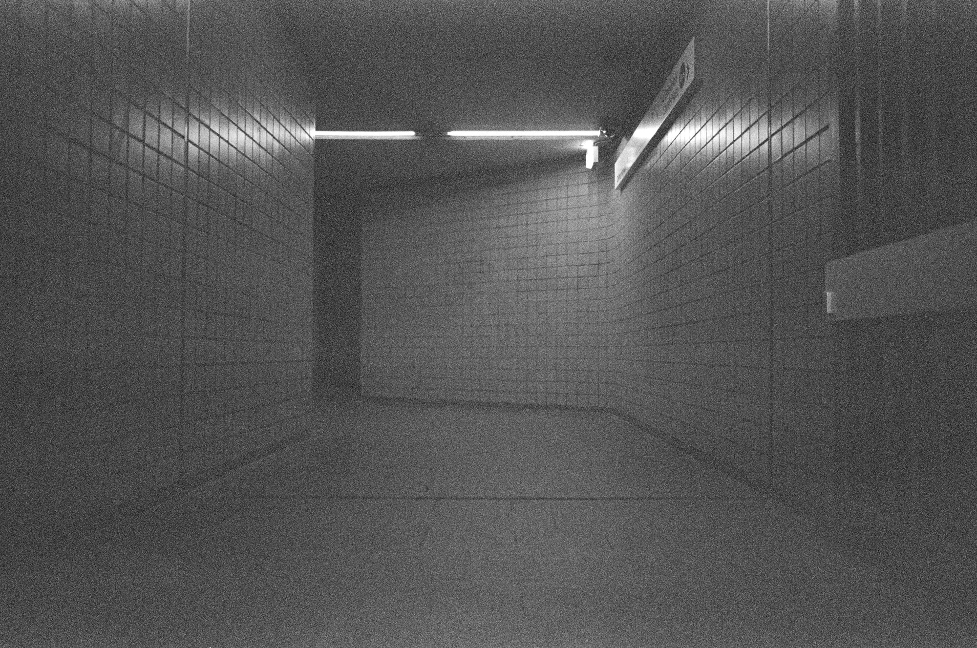 A tiled pedestrian underpass ending in a blank wall, lit by a single fluorescent tube on the ceiling, with nobody in it